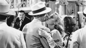 A black and white image shows a woman in a white glove intimately leaning into a man with a hat, surrounded by other people in formal attire at a vintage-style venue.