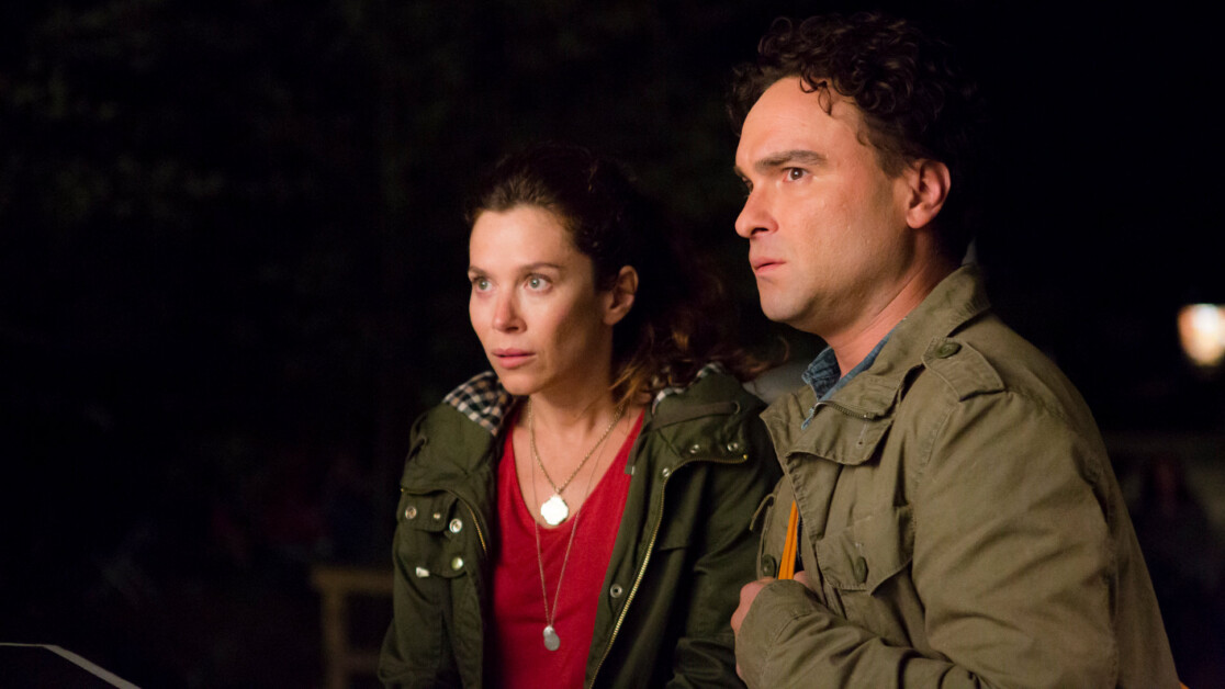 A woman and a man, both wearing jackets, stand outside at night, looking forward with serious expressions.