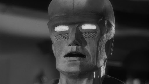 Close-up of a humanoid robot with illuminated rectangular eyes, a smooth metallic face, and an expressionless mouth, depicted in black and white.