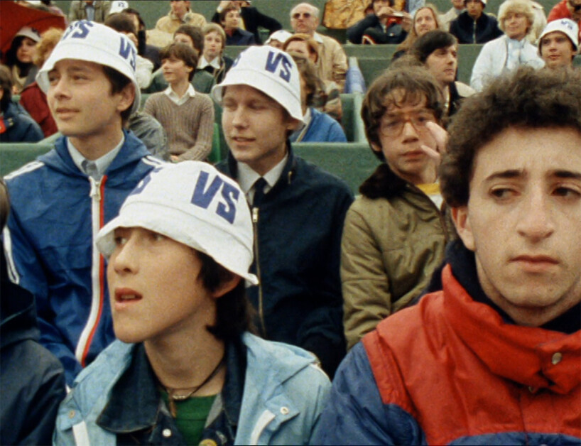 A group of people, mostly young men, are seated in a stadium. Several wear white hats labeled "VS." Some look attentive while others appear bored. The crowd in the background is also watching.