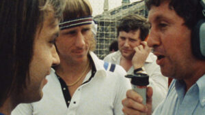 ‘The French’ Blu-Ray Review – A Revealing Look At Historic Sports Event Four men are interacting at what appears to be an outdoor event. One man, holding a microphone with a foam cover, seems to be conducting an interview. Another wears a headband and listens intently.