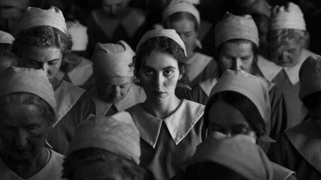 A group of women in uniform, most with heads bowed, stand in rows. One woman in the center looks directly at the camera. The image is in black and white.