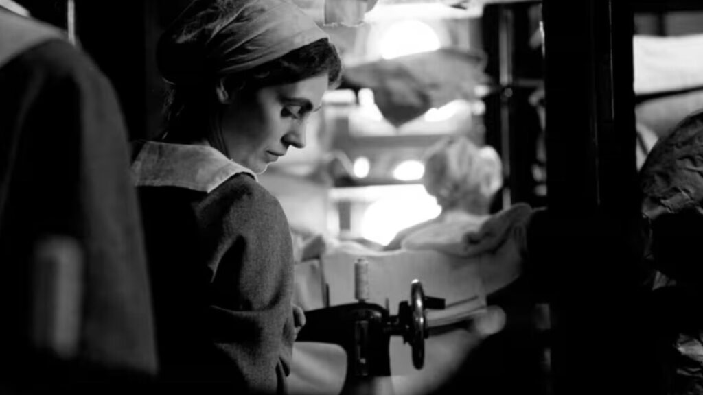 Black and white image of a woman with a headscarf standing next to a piece of equipment, appearing to work with fabric or materials. Other people and items are visible in the background.
