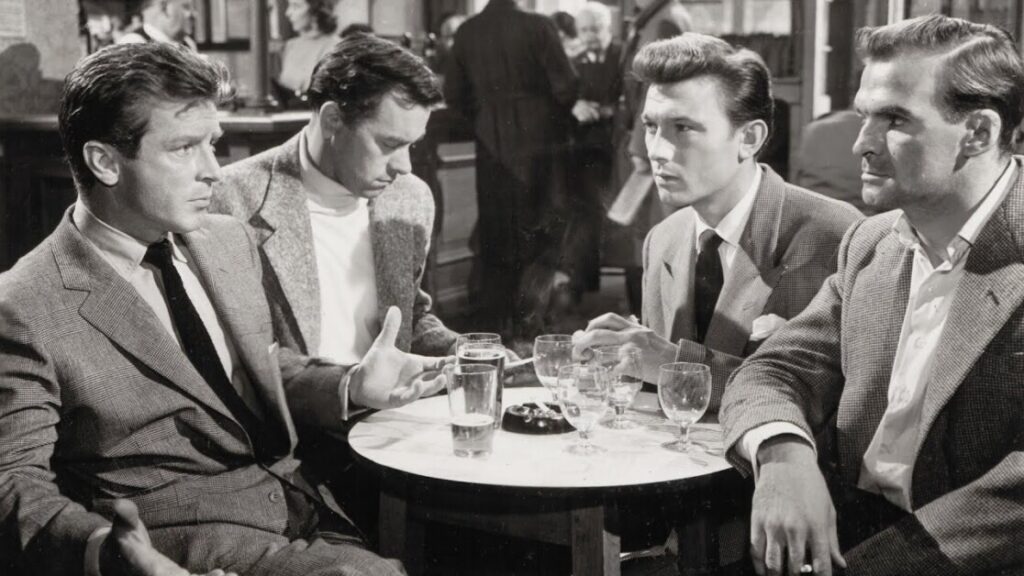 Four men in suits sit around a small round table with drinks in a busy bar, engaged in a serious conversation.