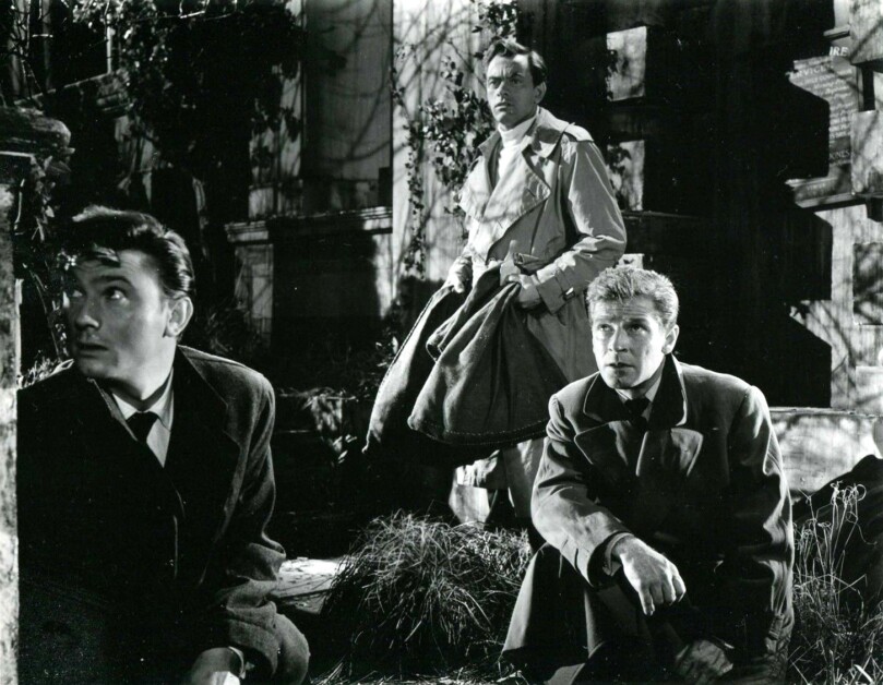 Three men, dressed in suits and coats, are positioned near a building's entrance; one kneels in the foreground, while another stands holding a bag, and the third man looks apprehensively to the side.