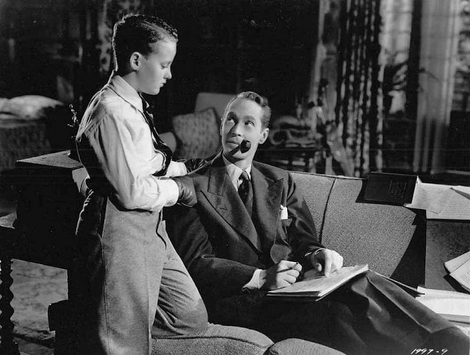 A man with a pipe sits on a couch, holding a book and pen, while a young boy in a sweater and tie stands beside him, looking at the man.