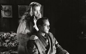 ‘The Hour Before The Dawn’ Blu-Ray Review – Sinister WWII Drama Is An Engrossing Watch A woman rests her hand on the shoulder of a seated man in a room with framed photos on the wall. Both appear deep in thought.