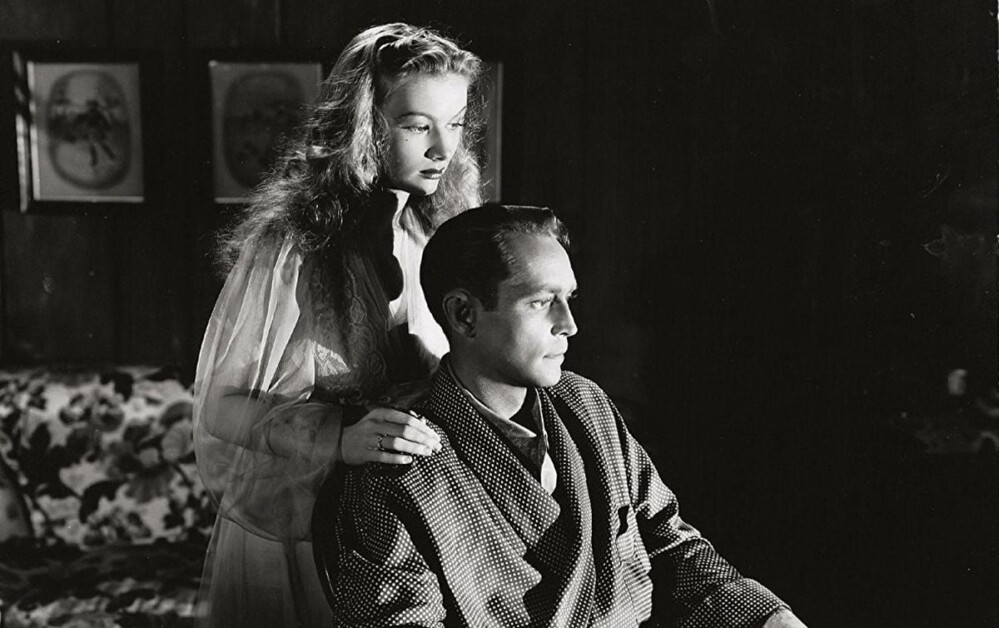 A woman rests her hand on the shoulder of a seated man in a room with framed photos on the wall. Both appear deep in thought.