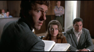 ‘The Lawyer’ (1970) Blu-Ray Review – Straightforward Legal Drama Provides Decent Entertainment A man in a grey suit looks intently to the side in a courtroom. A woman and another man sit behind him, with a police officer standing guard near the wall.
