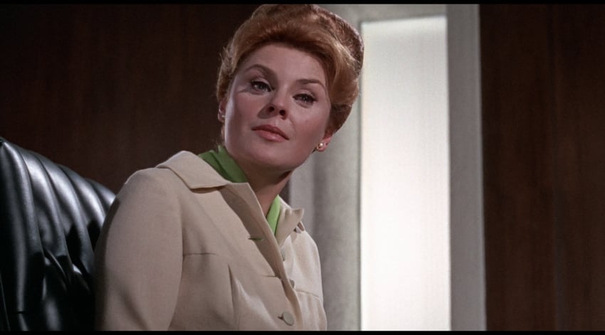 A person with an updo hairstyle and a beige jacket sits in a dark-colored chair, looking slightly to the side.