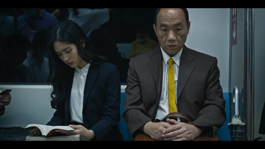 A young woman reads a book while an older man with a yellow tie sits next to her on a subway train.
