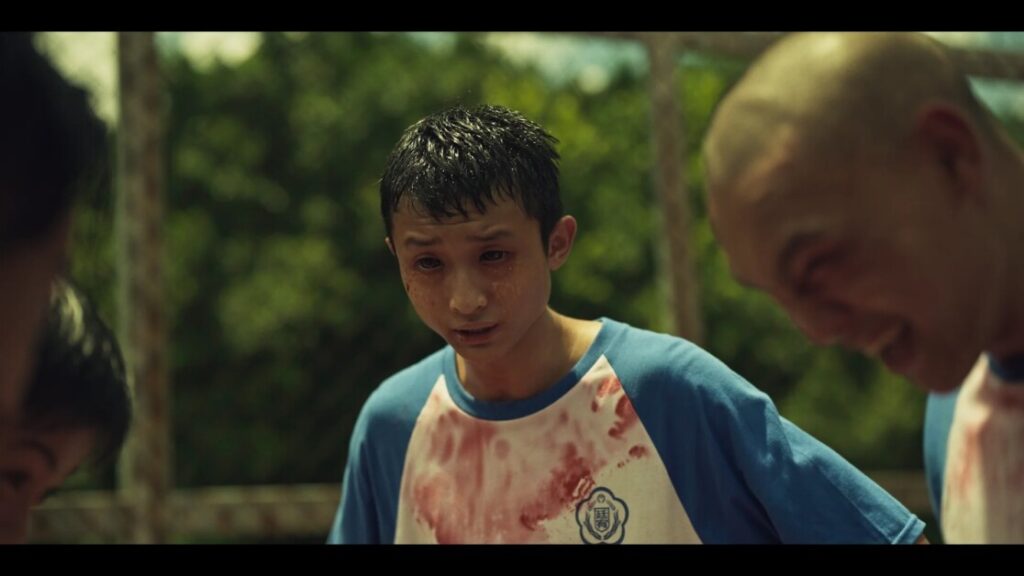 A young man in a blue and white shirt, drenched in sweat and with a bloodstain on his chest, stands outdoors surrounded by others with similar attire.
