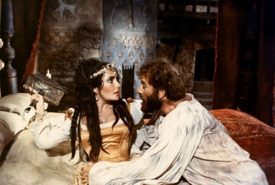 A woman in historical attire holds a book while lying on a bed, looking surprised at a man with a beard who is wearing a white shirt and leaning towards her. The background is a vintage room.