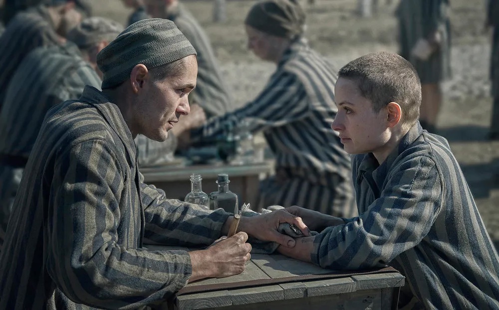 Two people in striped uniforms sit at a table, one giving a tattoo to the other. Other individuals in similar attire are visible in the background.