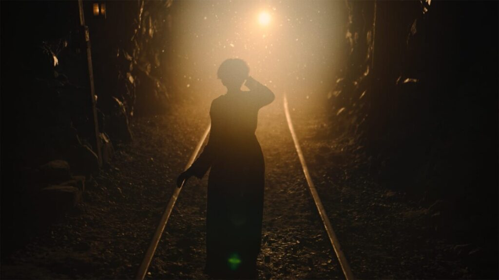A person stands on a railway track in a dimly lit tunnel, illuminated from behind by a bright light in the distance.