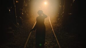 A person stands on a railway track in a dimly lit tunnel, illuminated from behind by a bright light in the distance.