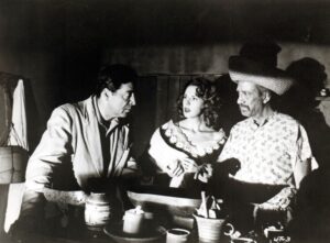 Sci-Fi Chillers Collection Blu-Ray Review – A Fun & Silly Blast From The Past Three people converse at a table in a dimly lit room. The man on the left wears a suit, the woman in the center has a shawl, and the man on the right wears a cowboy hat and patterned shirt.