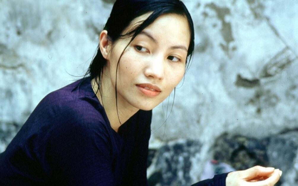 A woman with dark hair wearing a purple top is looking to the side while sitting against a textured wall background.