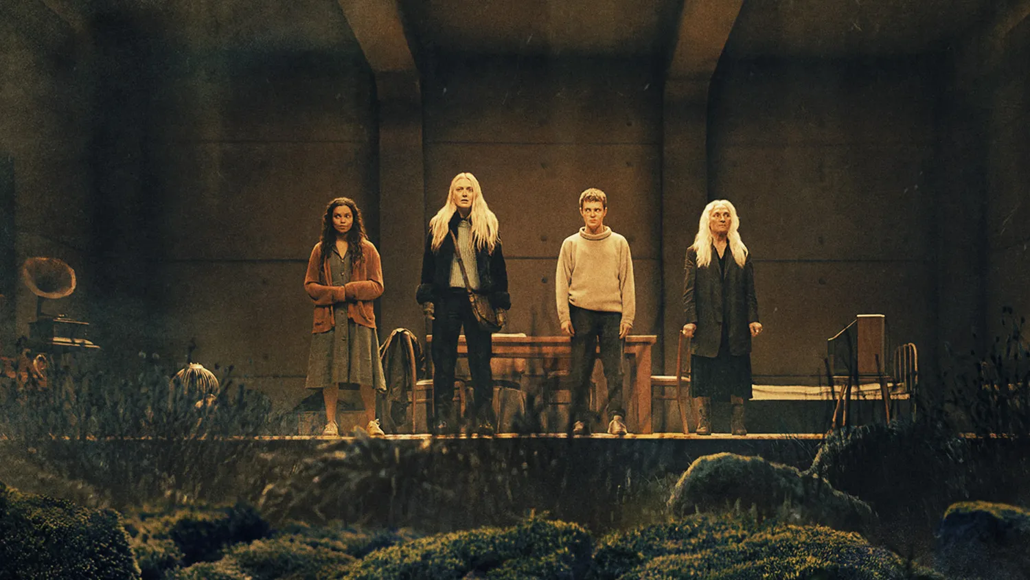 Four people stand side by side on a dimly lit stage inside an industrial-looking building. Two women are on the ends and two men are in the middle, all wearing casual clothes.