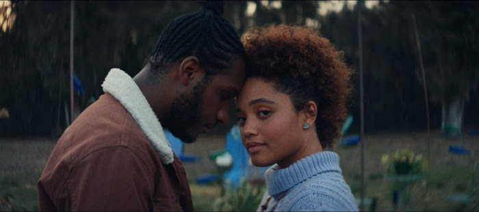 A man and woman stand closely with their foreheads touching outdoors, sharing an intimate moment with a soft expression.