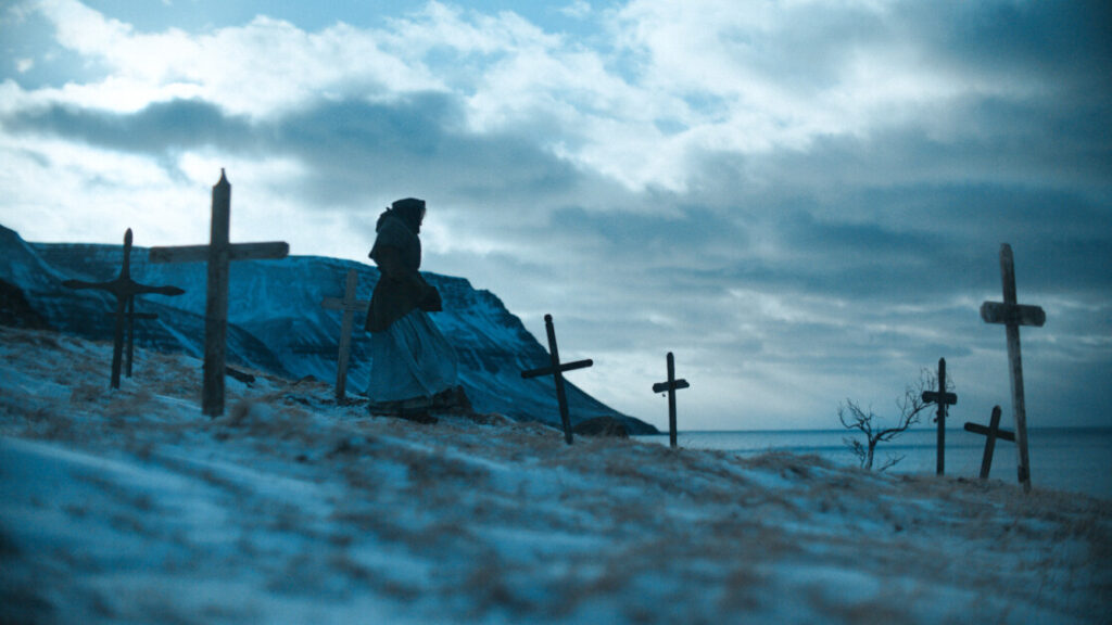 A person walks through a snow-covered landscape with wooden crosses, under a cloudy sky near a body of water and mountains in the background.
