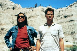 ‘Thunderheart’ Blu-Ray Review – A Thoughtful, Thrilling Native Drama Two men standing in front of a rocky landscape with onlookers on the cliffs above them. The man on the left wears a denim jacket and a red shirt, and the man on the right wears a white polo shirt.