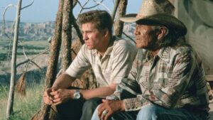 Two men sit outdoors; one is younger wearing a light shirt, the other is older wearing a checkered shirt and a straw cowboy hat. They are surrounded by nature with a scenic landscape in the background.