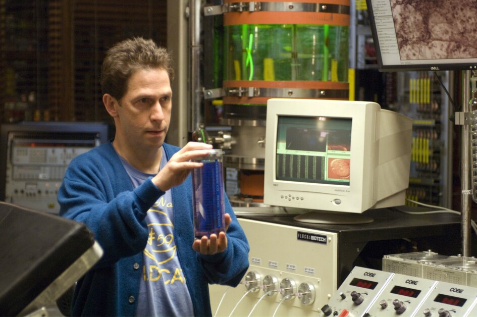 A person wearing a blue sweater holds a rectangular blue object in a laboratory filled with various scientific equipment, including monitors, buttons, and dials.