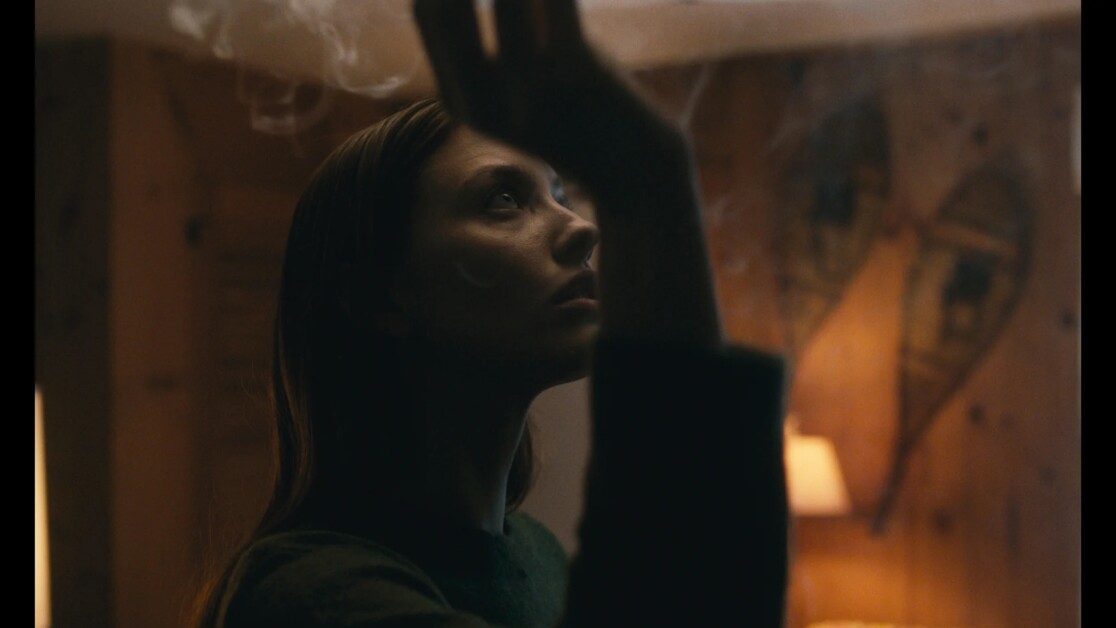 A person with long hair raises a hand, looking up pensively in a dimly lit room with wooden walls and snowshoes hanging as decor. Smoke or mist is visible around the hand.