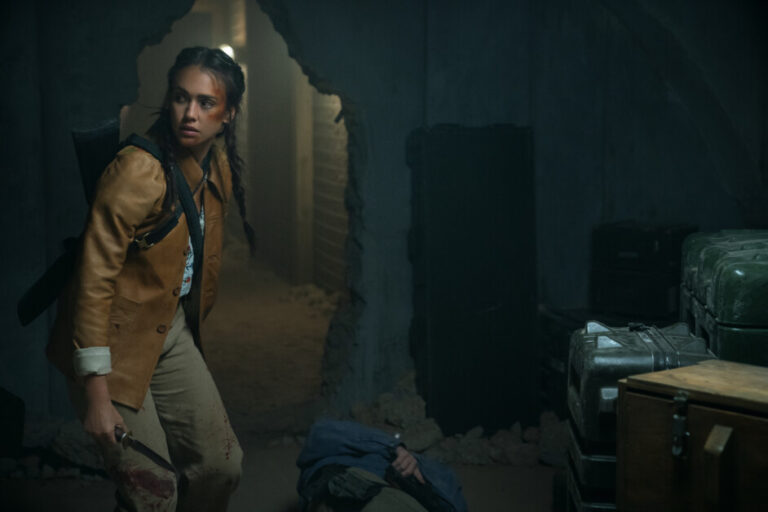 ‘Trigger Warning’ Review – Jessica Alba’s Most Badass Film Yet A woman carrying a knife and a backpack stands in a dimly lit, cluttered room with a large hole in the wall and a person lying on the ground nearby.