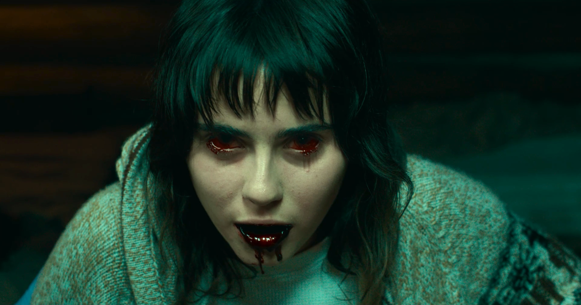 A person with long dark hair and glowing red eyes is shown with blood around their mouth and streams of blood flowing from their eyes. They are wearing a gray sweater.