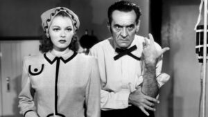 A woman and a man are standing side by side. The woman wears a headscarf and a tailored jacket, while the man has a stern expression and wears a bow tie, with his arm bent at the elbow.