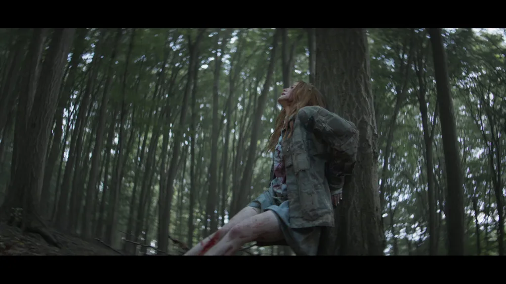 A person with injuries leans against a tree in a dense forest, looking upwards. They are wearing a jacket and shorts.
