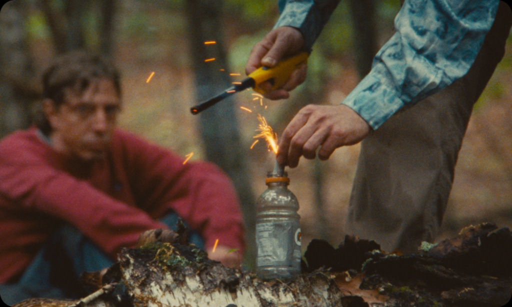 Two individuals outdoors. One person lights a sparkler with a lighter held above a Gatorade bottle on a tree stump, while another person sits in the background, watching.