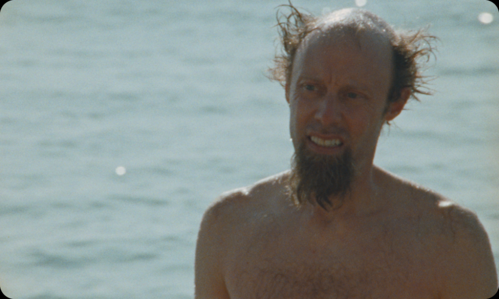 A shirtless man with disheveled hair and a goatee stands in front of a body of water.