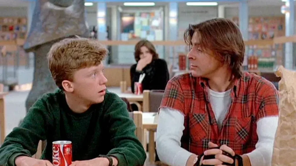 Two young men sit at a table with soda cans in front of them, engaged in conversation. Another person sits in the background, partially obscured.