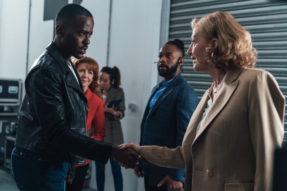 A man in a black leather jacket and a woman in a beige blazer shake hands in an industrial-looking room, with three other people observing in the background.
