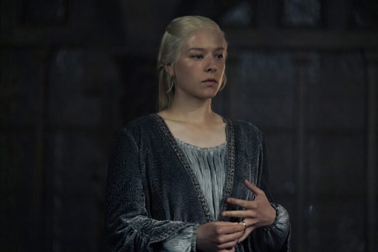 ‘House Of The Dragon’ Season 2 Episode 2 Review – The Fallout From Tragedy A person with long white hair, dressed in a dark robe, stands in a dimly lit room with hands positioned near their chest.