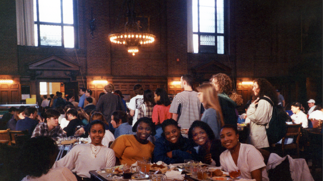 A group of five people smiling and seated at a table in a crowded dining hall, with other people standing and sitting in the background.
