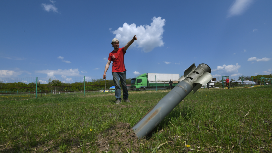 A person points towards a large missile lodged in the ground in a grassy area with a road and trucks visible in the background under a clear blue sky.