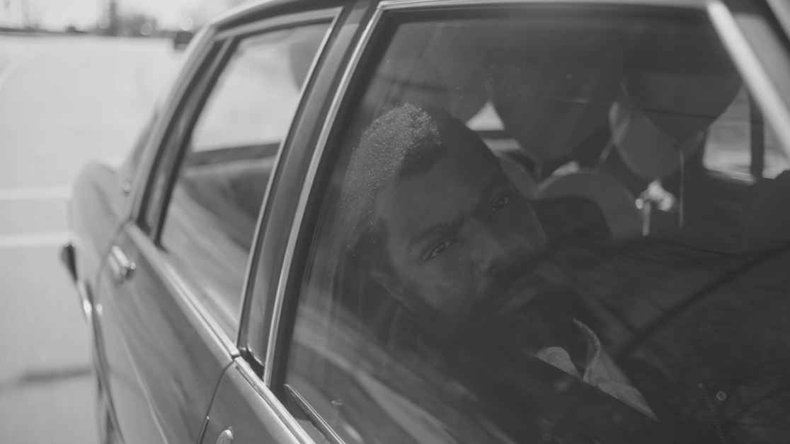 A man with a serious expression looks out from the back seat of a car window, captured in black and white.