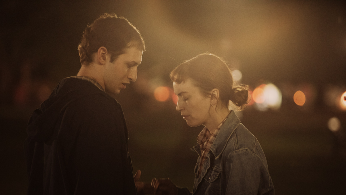 Two individuals stand close to each other at night in an outdoor setting with blurred lights in the background, appearing to engage in a serious conversation.