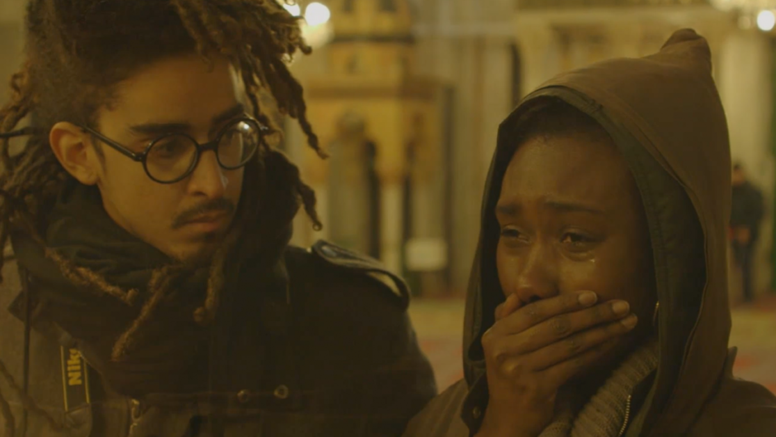 Two individuals are indoors, with one person wearing glasses and looking at the other, who is crying and covering their mouth with a hand.