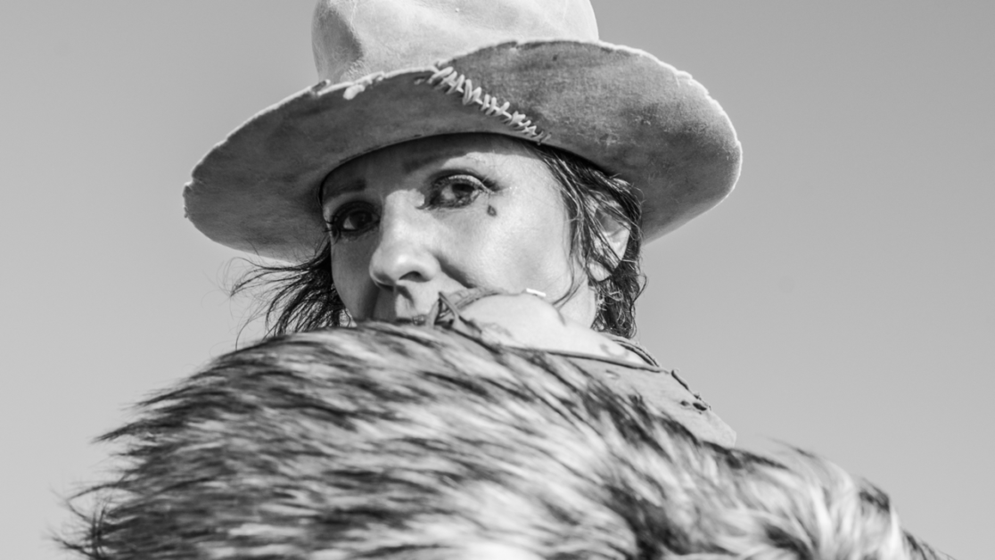 Black and white photo of a person wearing a worn hat and holding a fur-covered item, gazing into the distance.