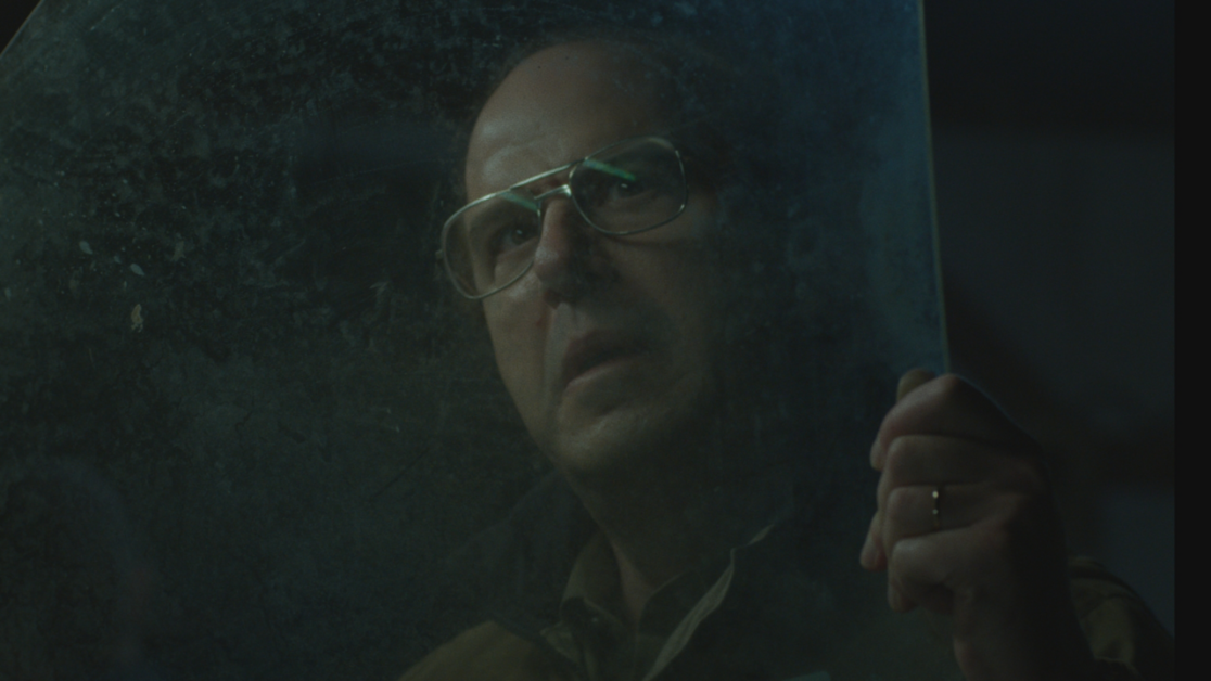 A man wearing glasses holds up and examines a semi-transparent, dusty sheet of glass or plastic, with a focused expression.
