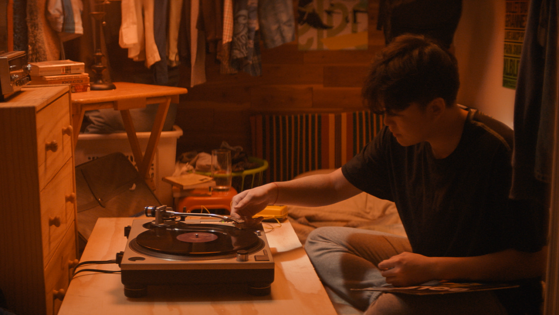 A person sits on the floor of a dimly lit room, operating a turntable. The room has a dresser, a desk, clothes hanging, and various items scattered around.