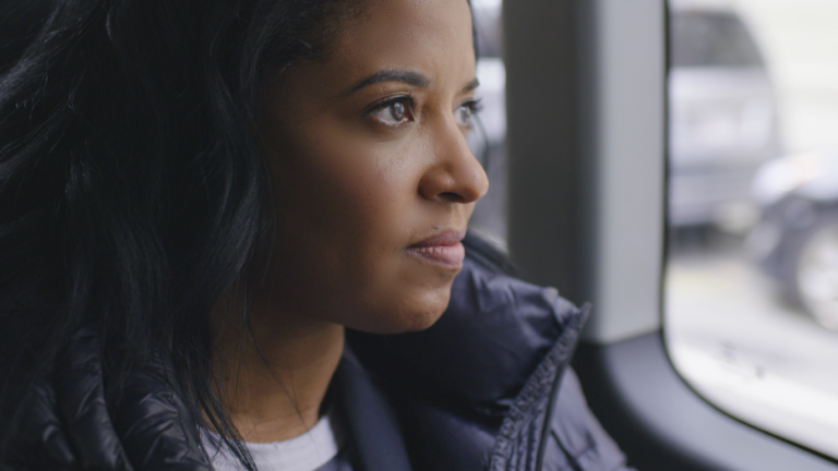 A person with long hair wearing a puffer jacket looks out of a window.