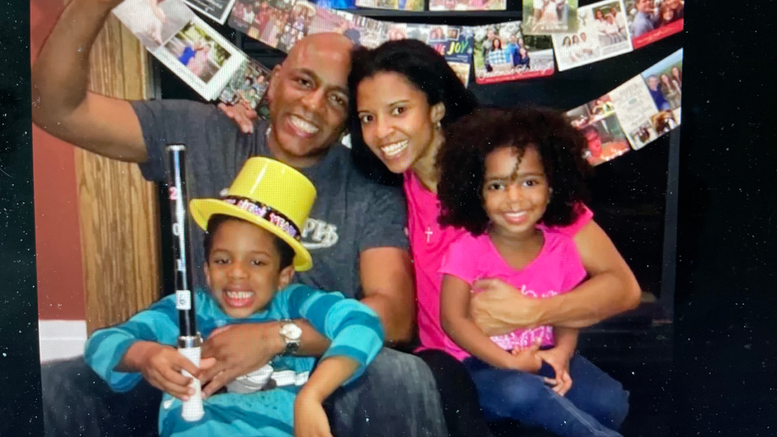 A smiling family of four poses indoors. The father holds a toy sword while the son wears a yellow hat. The mother embraces the daughter. Greeting cards are displayed in the background.