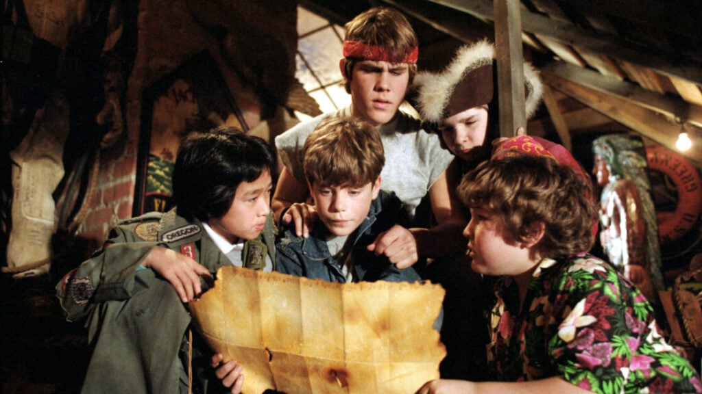 Five children are huddled together in an attic, closely examining an old, yellowed map. Various items and wooden beams are visible in the background.