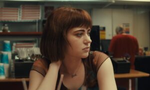 A woman with brown hair sits in a diner with a thoughtful expression, resting her hand on the back of her neck. A person in a red shirt is in the background.